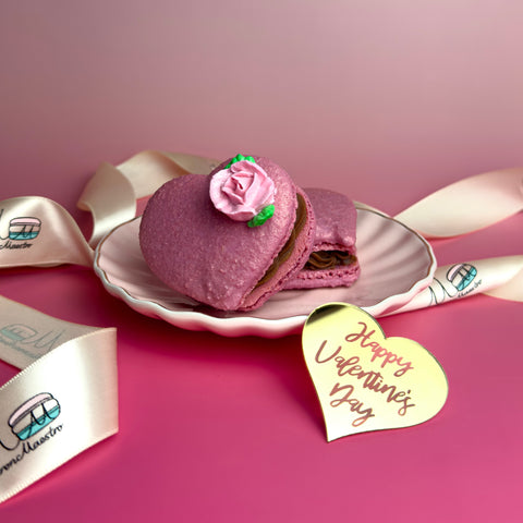 Pink macaron with a rose on a white plate, surrounded by ribbons and a 'Happy Valentine's Day' card on a pink background.